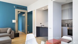 Een moderne woonkamer met een bank, een glazen salontafel en een keukenhoek met een fornuis en kasten.