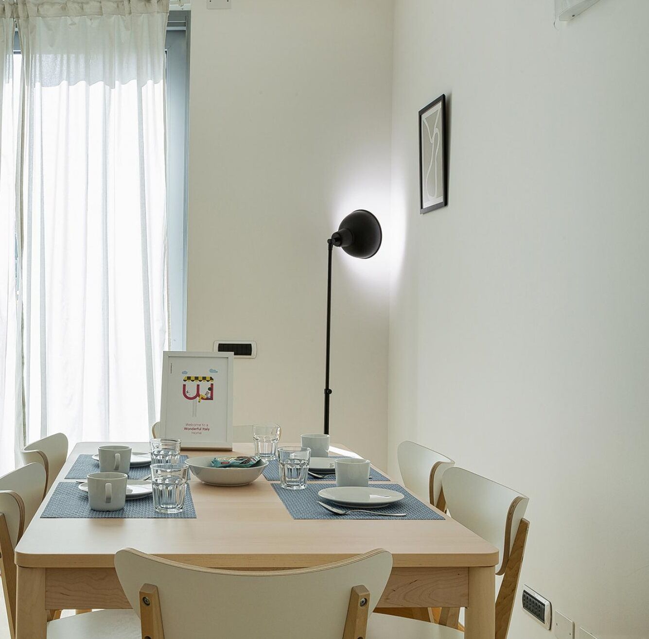 Uma sala de jantar com uma mesa posta para dois, um abajur preto e um quadro na parede.