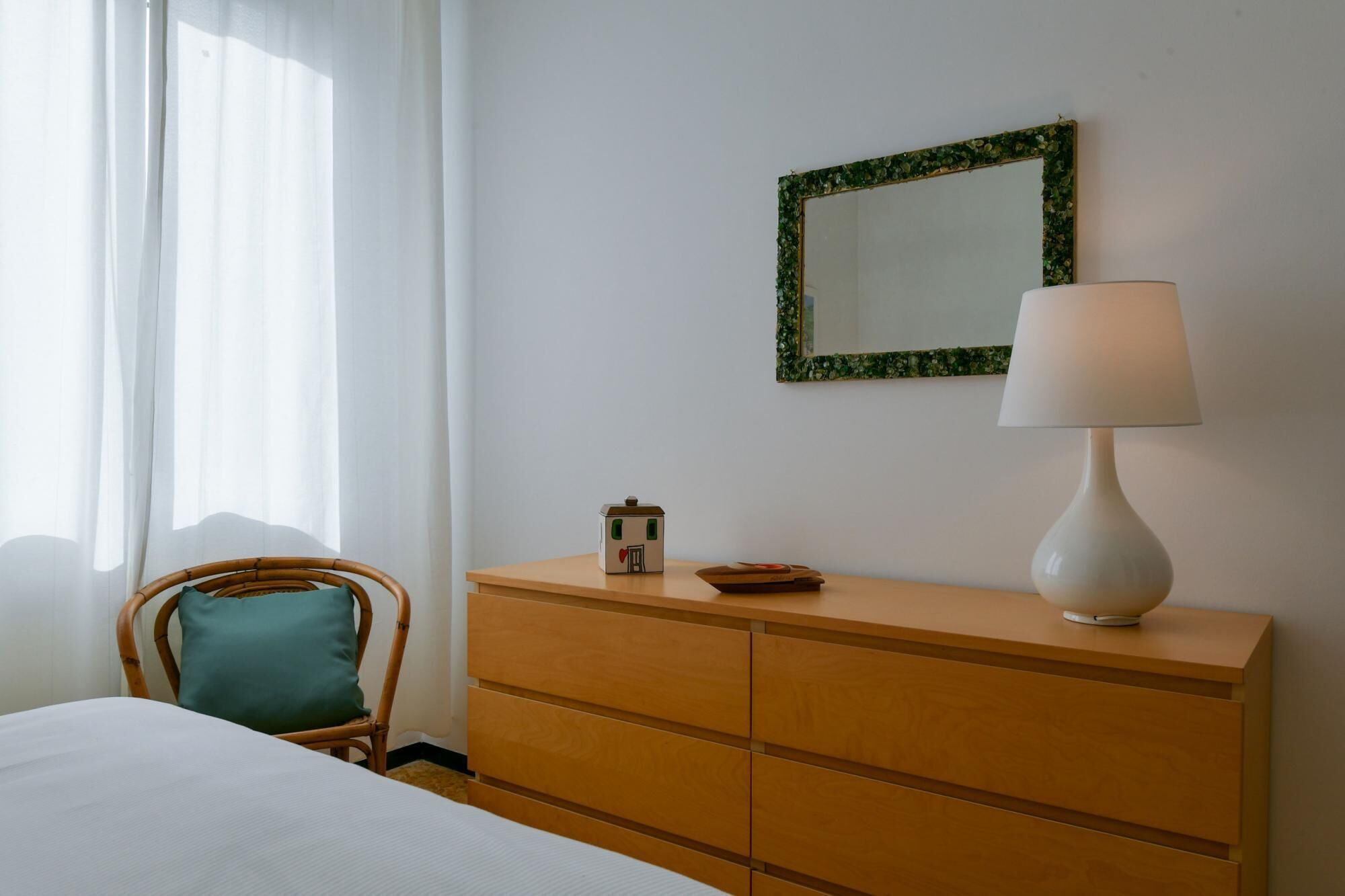 A neatly arranged bedroom with a bed, a wooden dresser, a mirror with a green frame, and a bedside lamp.