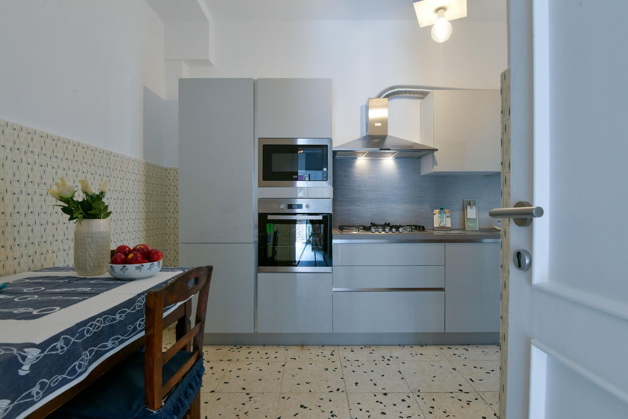 Une cuisine compacte équipée d’un four encastré, d’un four à micro-ondes et d’une cuisinière à gaz. On y trouve une table à manger agrémentée d’un vase de fleurs et d’un saladier de fruits. Le sol est recouvert de carreaux aux motifs pointillés.