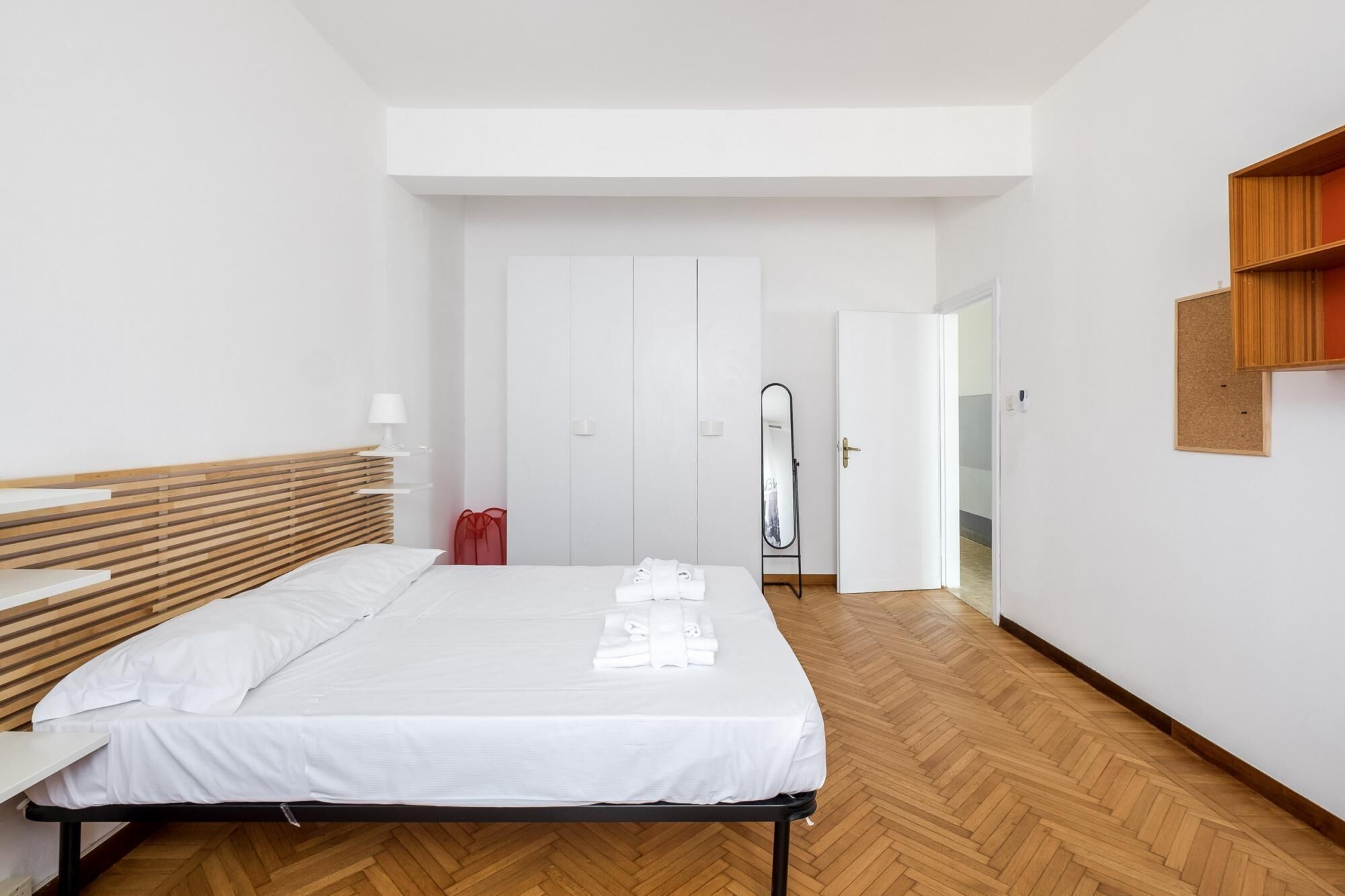 A modern hotel room with a wooden bed, white linens, a small bedside table, a mirror, and a wooden shelf.