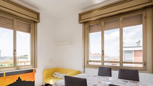 Ein Zimmer mit zwei großen Fenstern, einem gelben Sofa, einem Esstisch mit vier Stühlen und Blick auf Gebäude.