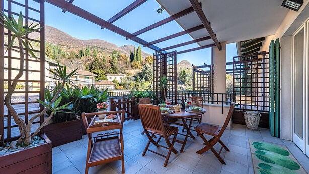 A covered patio with wooden furniture, potted plants, and a view of a mountainous landscape. - Al Vittoriale 3 by Wonderful Italy (Gardone Riviera)