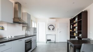 A modern kitchen with grey cabinetry, a built-in oven, and a stainless steel range hood. There is a dining area with a dark wooden table and chairs, and a bookshelf filled with books.