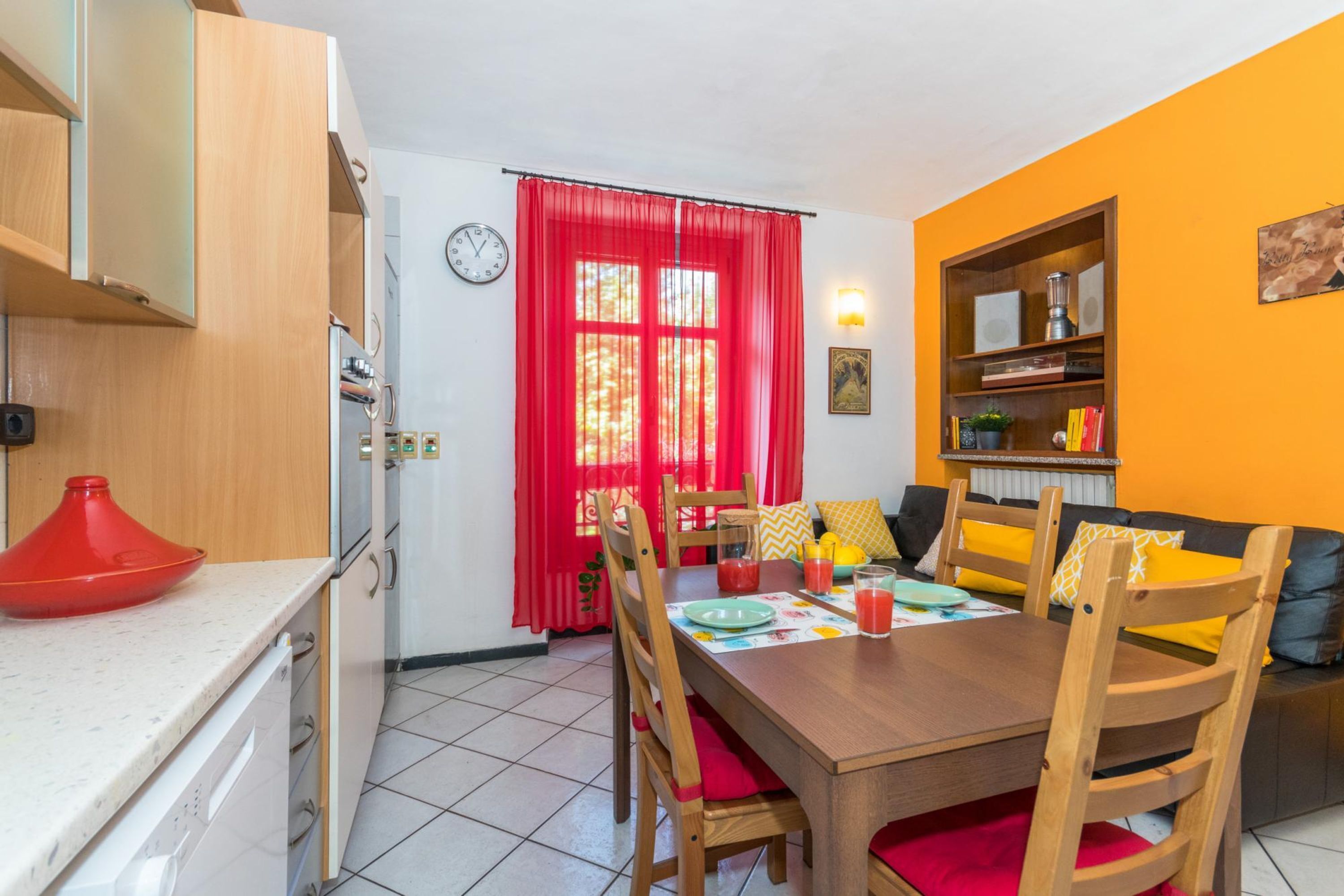 A compact kitchen with a dining area, wooden cabinets, a red curtain, and a bookshelf.