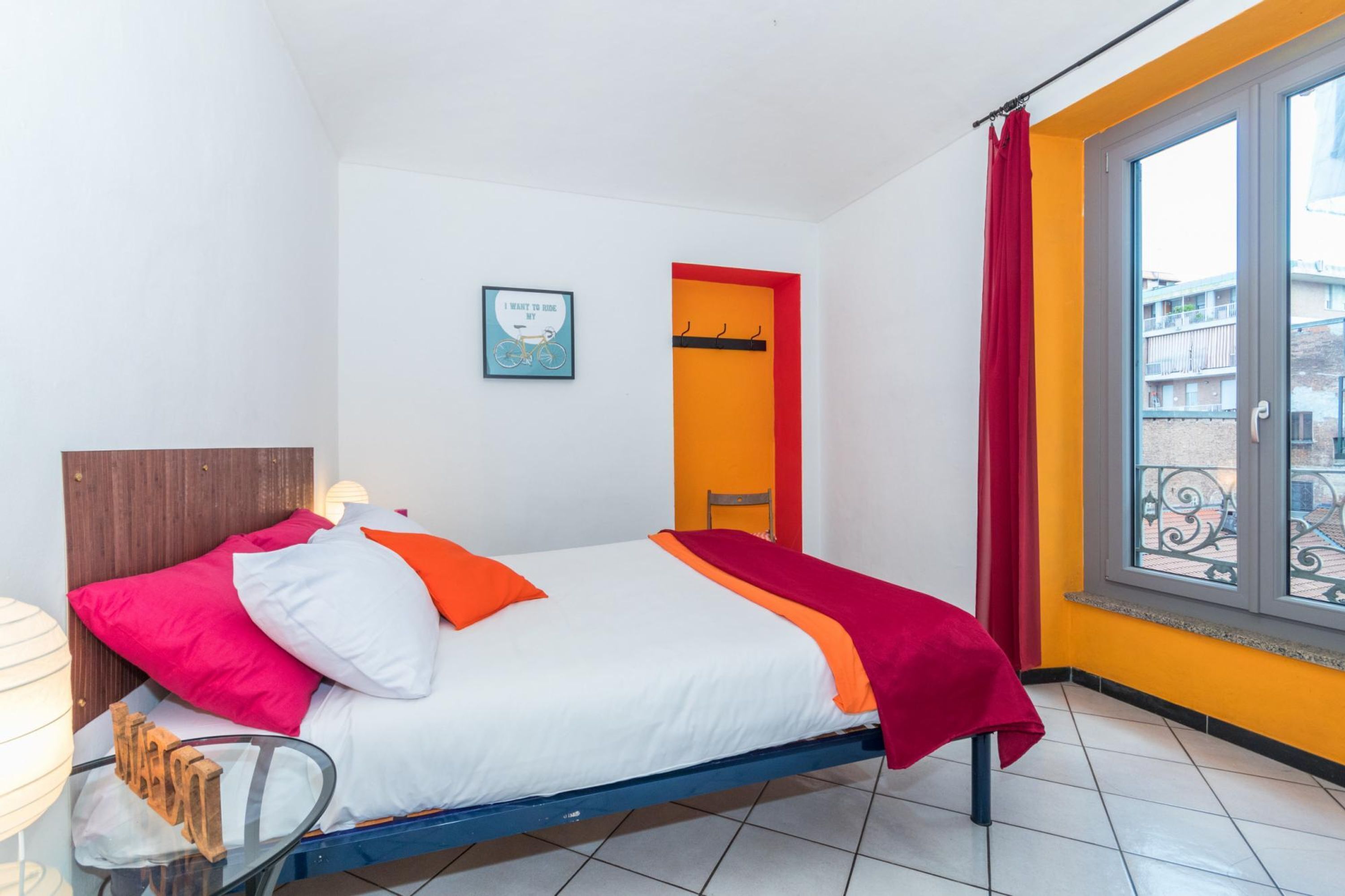 Een bed met een wit-oranje sprei, een rode deken en een houten hoofdbord. Aan de muur hangt een ingelijst foto, er is een raam met een rode gordijn en een balkon met een leuning.