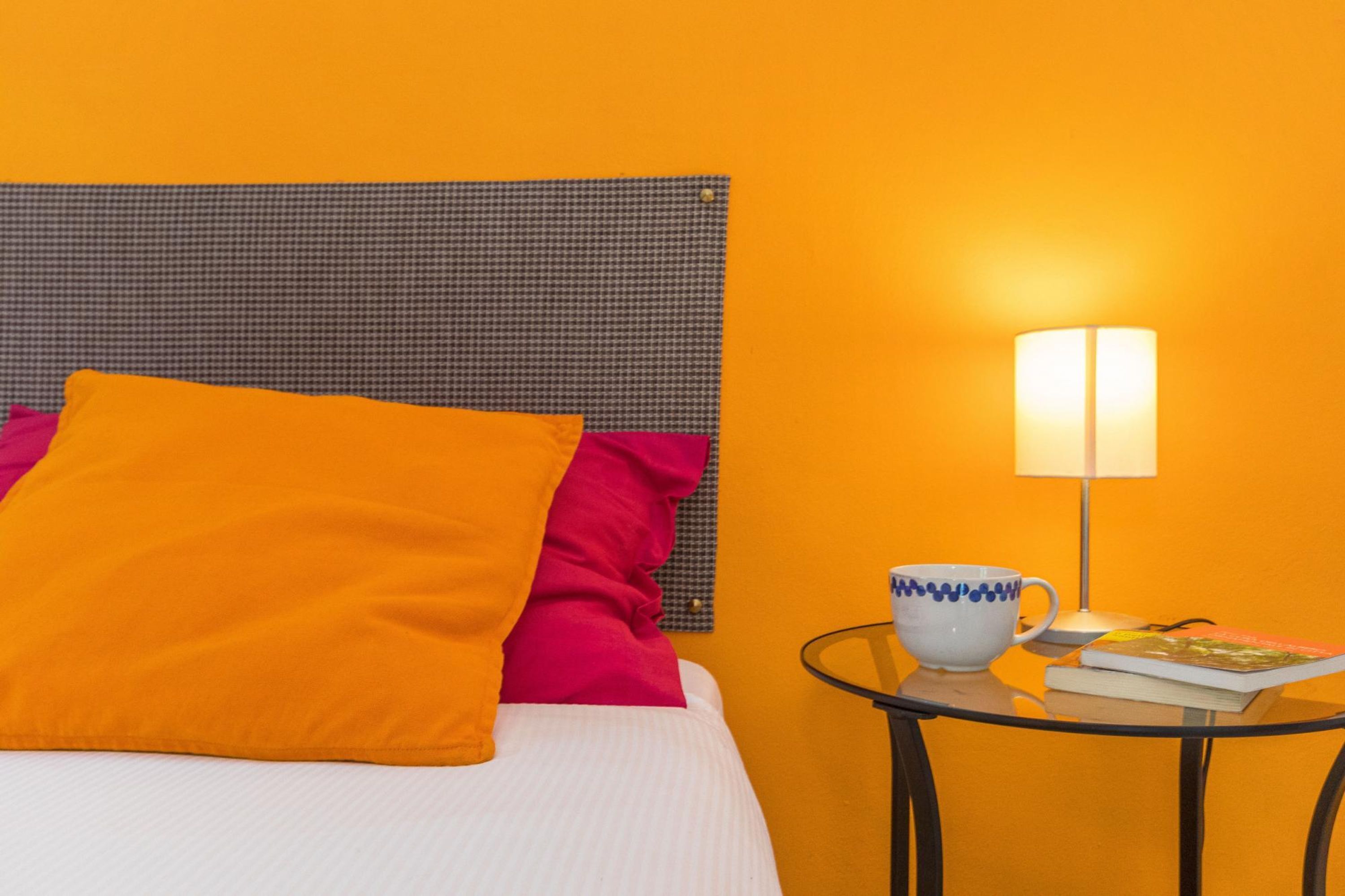 A bed with a checkered headboard, orange and pink pillows, a white bedspread, a glass table with a white mug, a book, and a lamp.