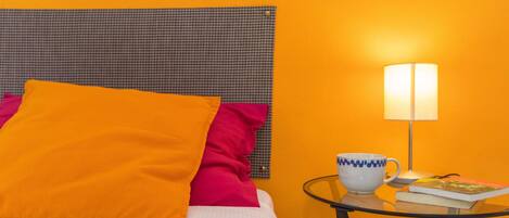 A bed with a checkered headboard, orange and pink pillows, a white bedspread, a glass table with a white mug, a book, and a lamp.