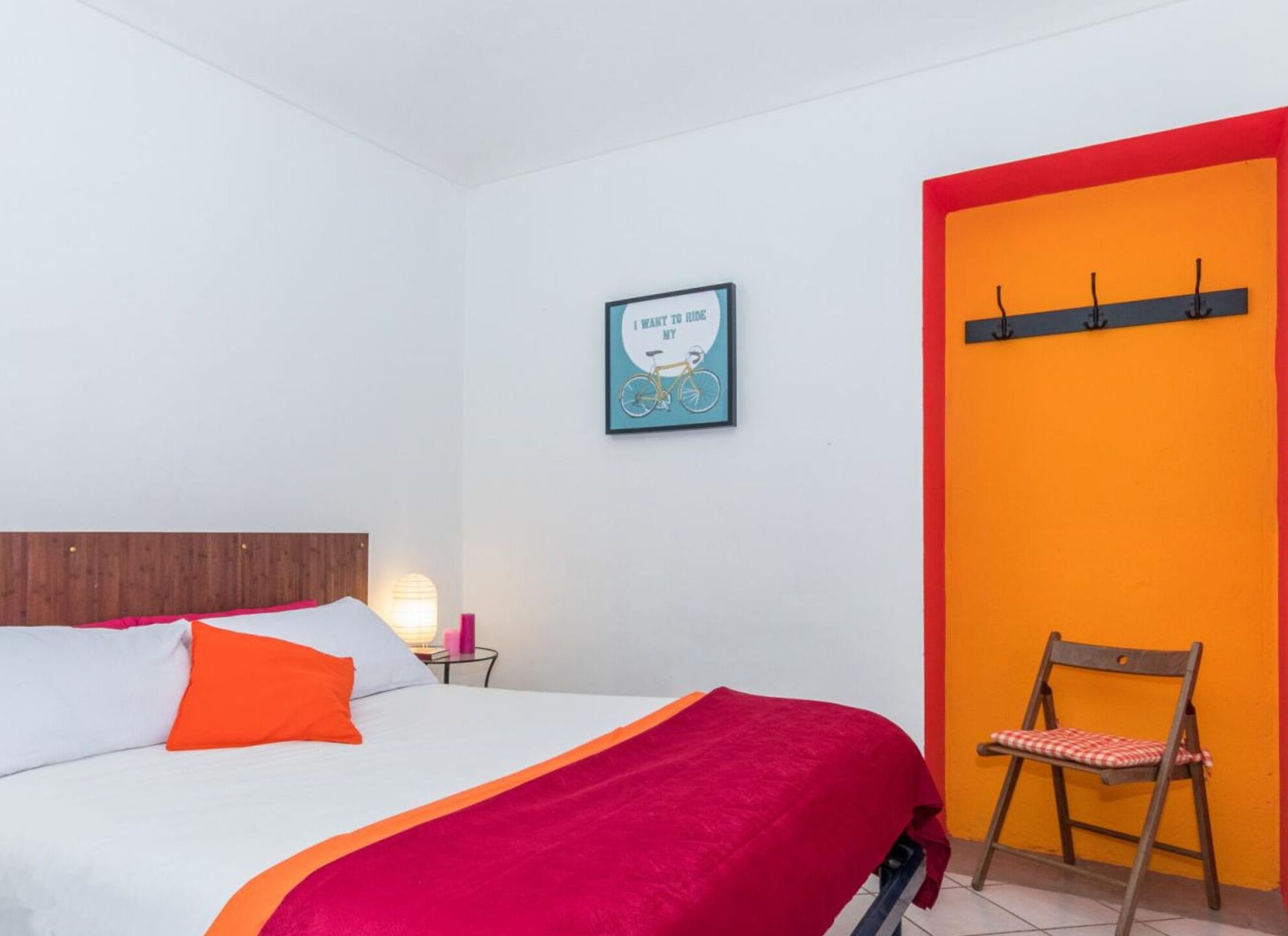 Een slaapkamer met een bed, nachtkastje, lamp, lijst met een poster en een stoel.