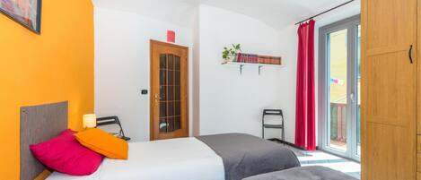 A bedroom with two beds, a wooden wardrobe, a window with red curtains, and a shelf with books and plants.
