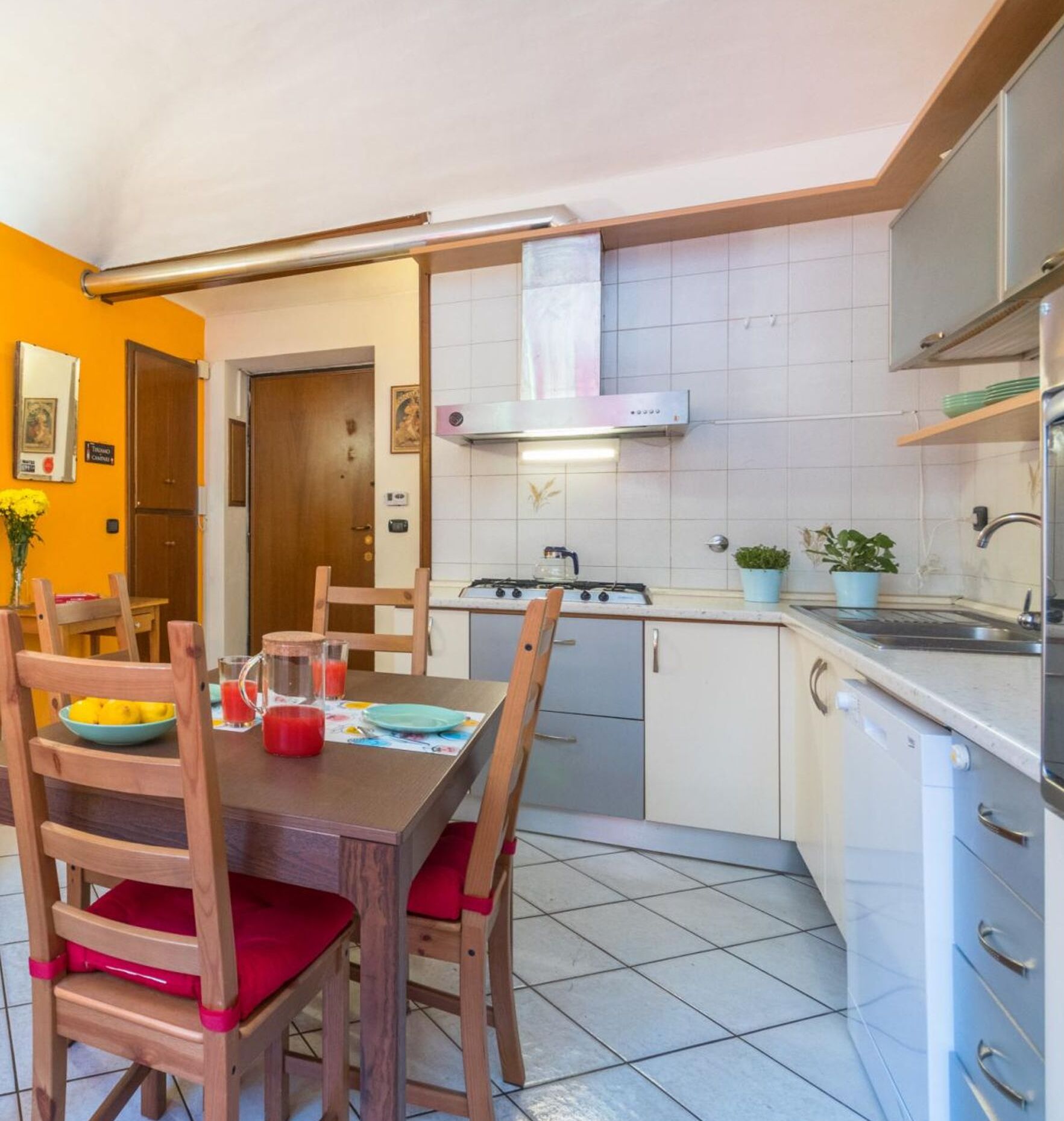 A compact kitchen with a gas stove, a sink, and a stove hood. There is a dining table with chairs and a vase of flowers.
