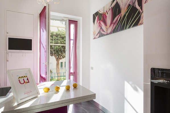 A kitchen with a countertop, a framed welcome sign, a window with pink curtains, and a painting on the wall.