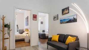 A modern apartment with a grey sofa, yellow cushions, a wooden coat rack, and a glass table. There are two beds with white bedding and a red "Thank you for not smoking" sign. - Torino 3 0 Sulla Via del Treno by Wonderful Italy (Turin)