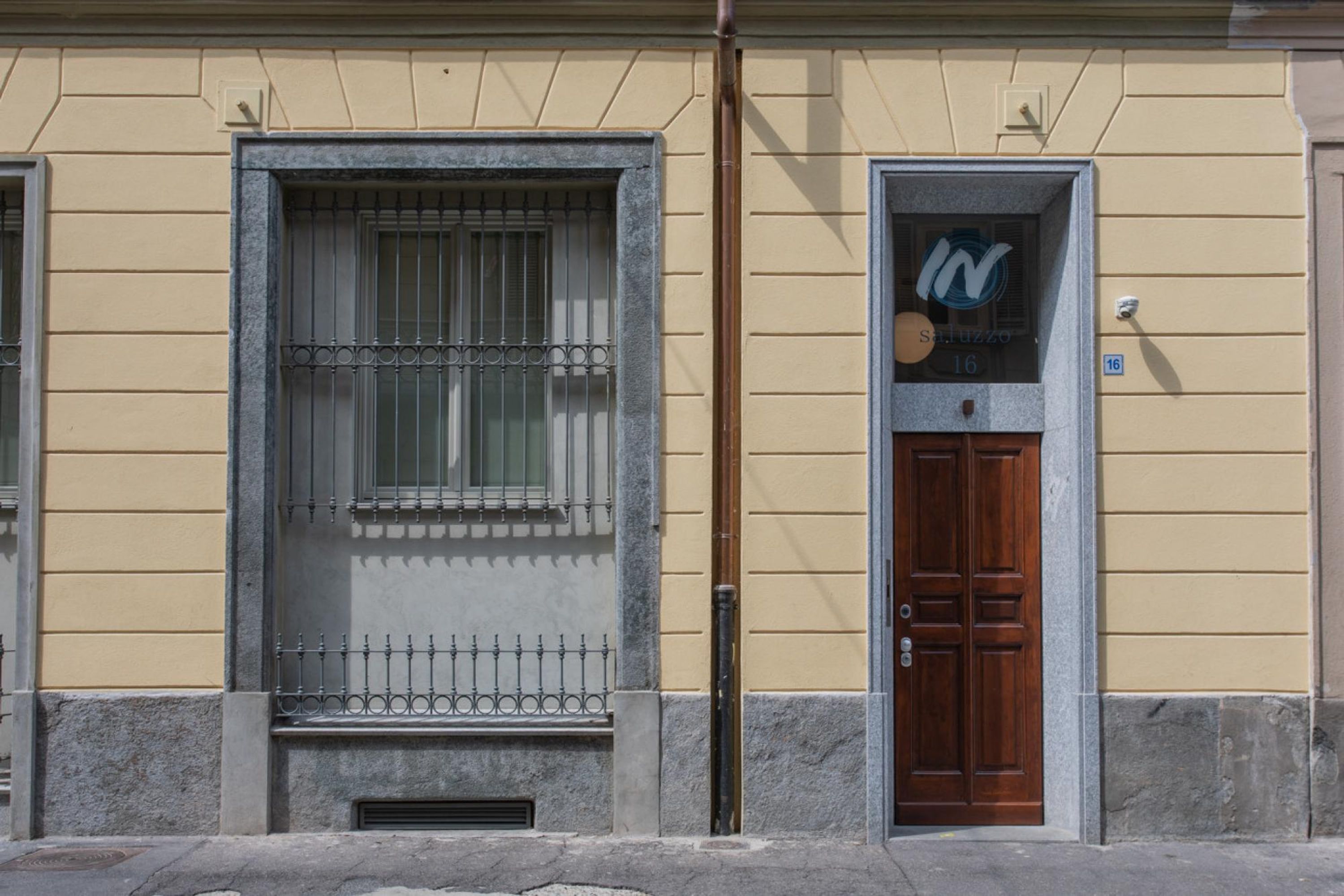 Fachada de edificio con una ventana protegida por barrotes de metal, una puerta de madera con un letrero encima y un ventanal con un emblema circular.
