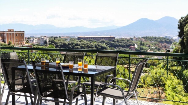 Une table entourée de chaises sur un balcon donnant sur une vue panoramique de la ville, avec des montagnes à l’horizon.