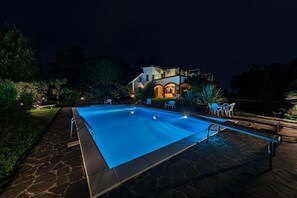 Une piscine avec une terrasse en pierre, une maison dont la façade est illuminée et du mobilier d’extérieur.