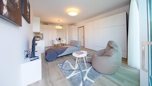 Un salon moderne avec un canapé, un fauteuil poire et une petite table. On y trouve un téléviseur à écran plat posé sur un meuble, une table basse et un tapis. La pièce est décorée dans des tons neutres et possède une grande fenêtre.