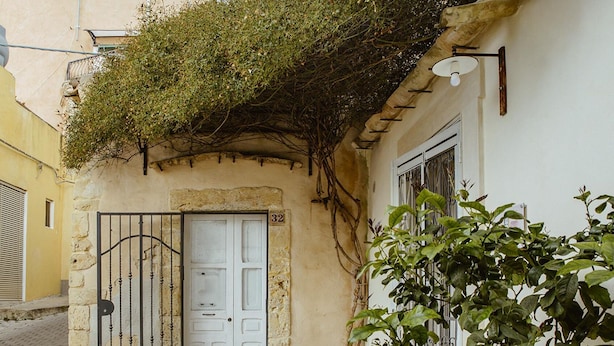 Una calle estrecha con un edificio amarillo, una puerta blanca y un portón metálico, y una planta al frente.