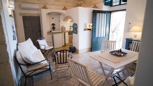 Un coin repas accueillant avec une table dressée pour deux personnes, une cuisine avec une porte bleue et un espace détente avec des chaises et des coussins.