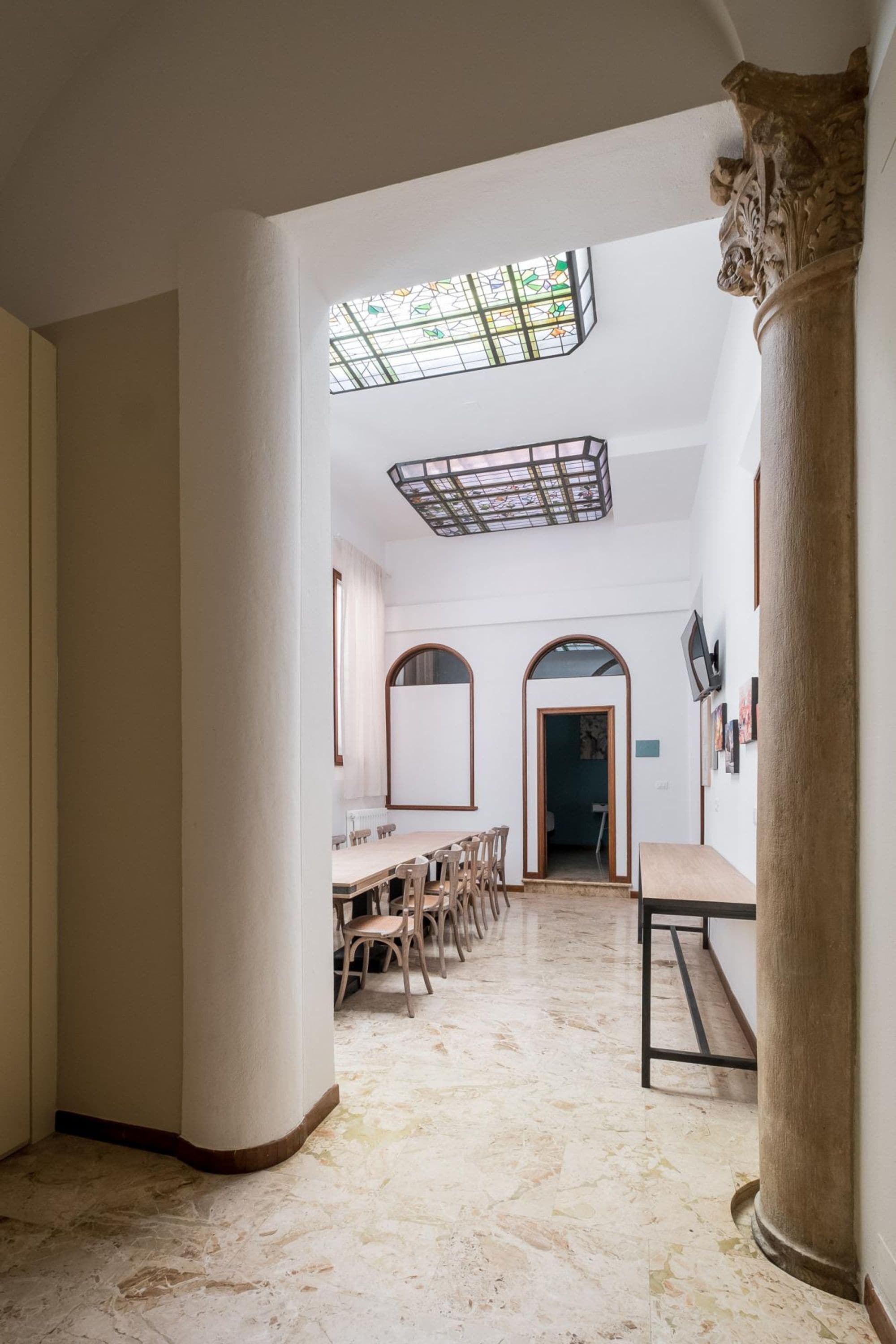 A spacious interior with a high ceiling, a dining area with wooden chairs, a large column, and a stained glass window.