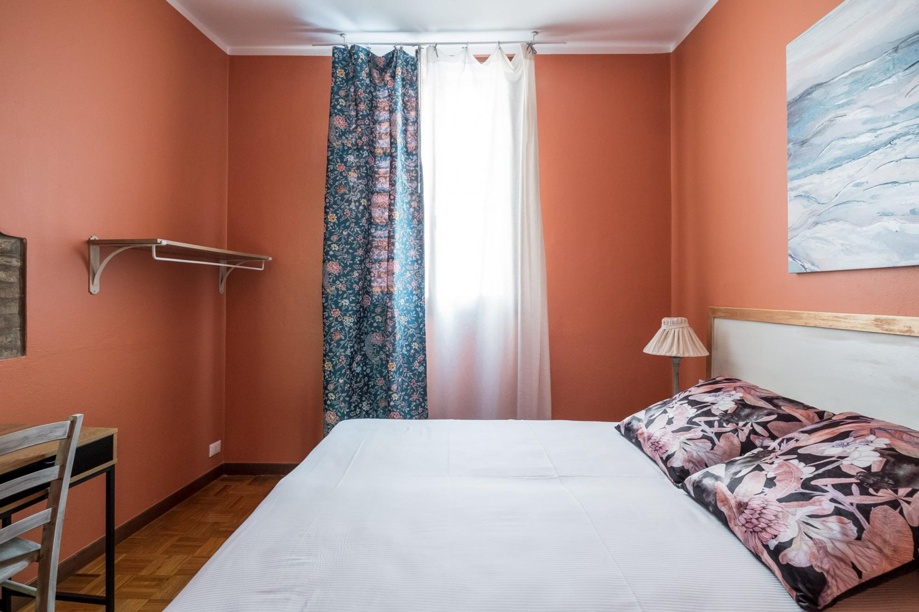 A bedroom with a bed, a wooden chair, a hanging shelf, a window with curtains, and a lamp.