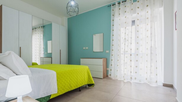 Un dormitorio con cama, mesita de noche, lámpara, cómoda, espejo y ventana con cortinas.