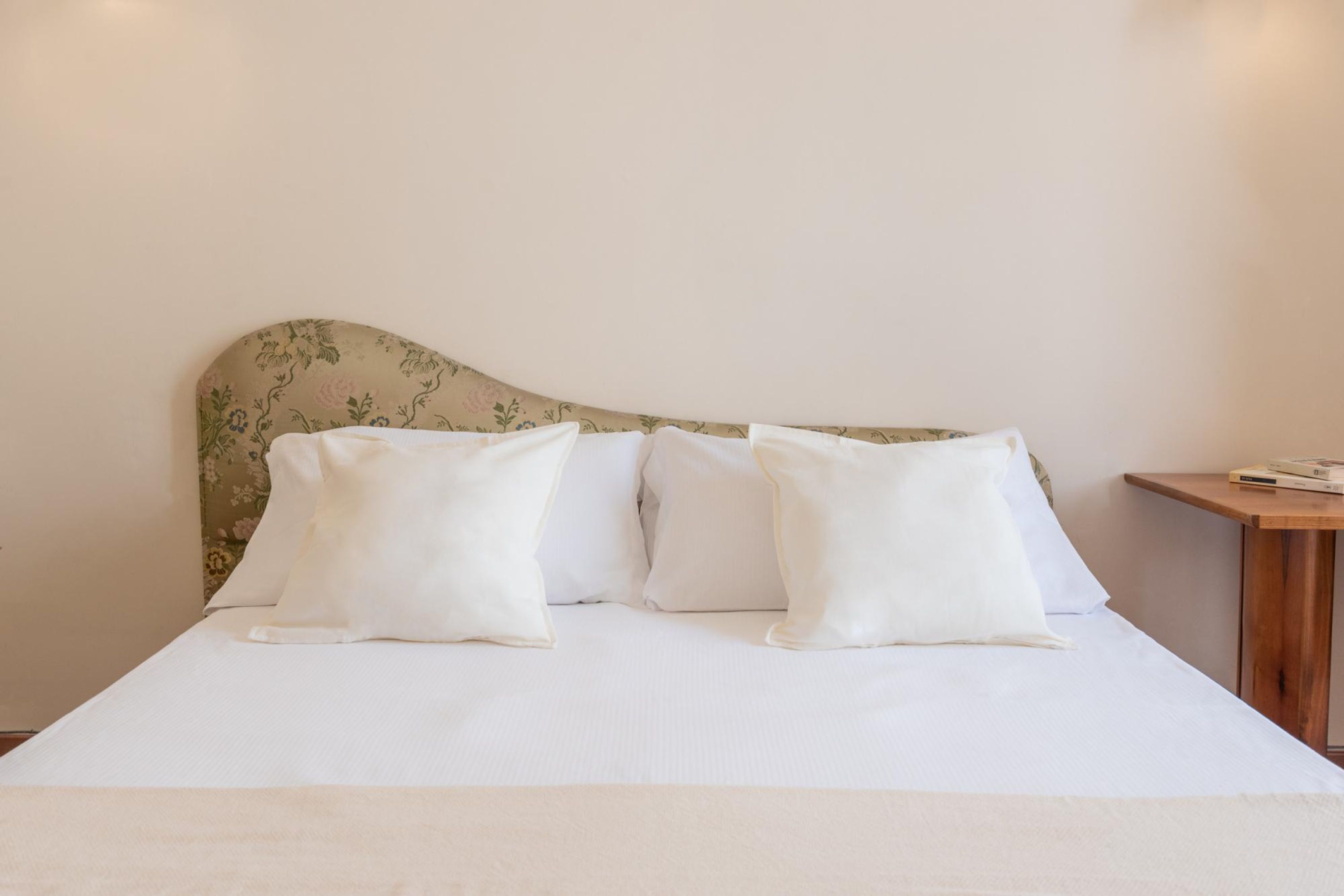 A neatly made bed with white linens and pillows, a wooden nightstand with a book, and a floral-patterned headboard.