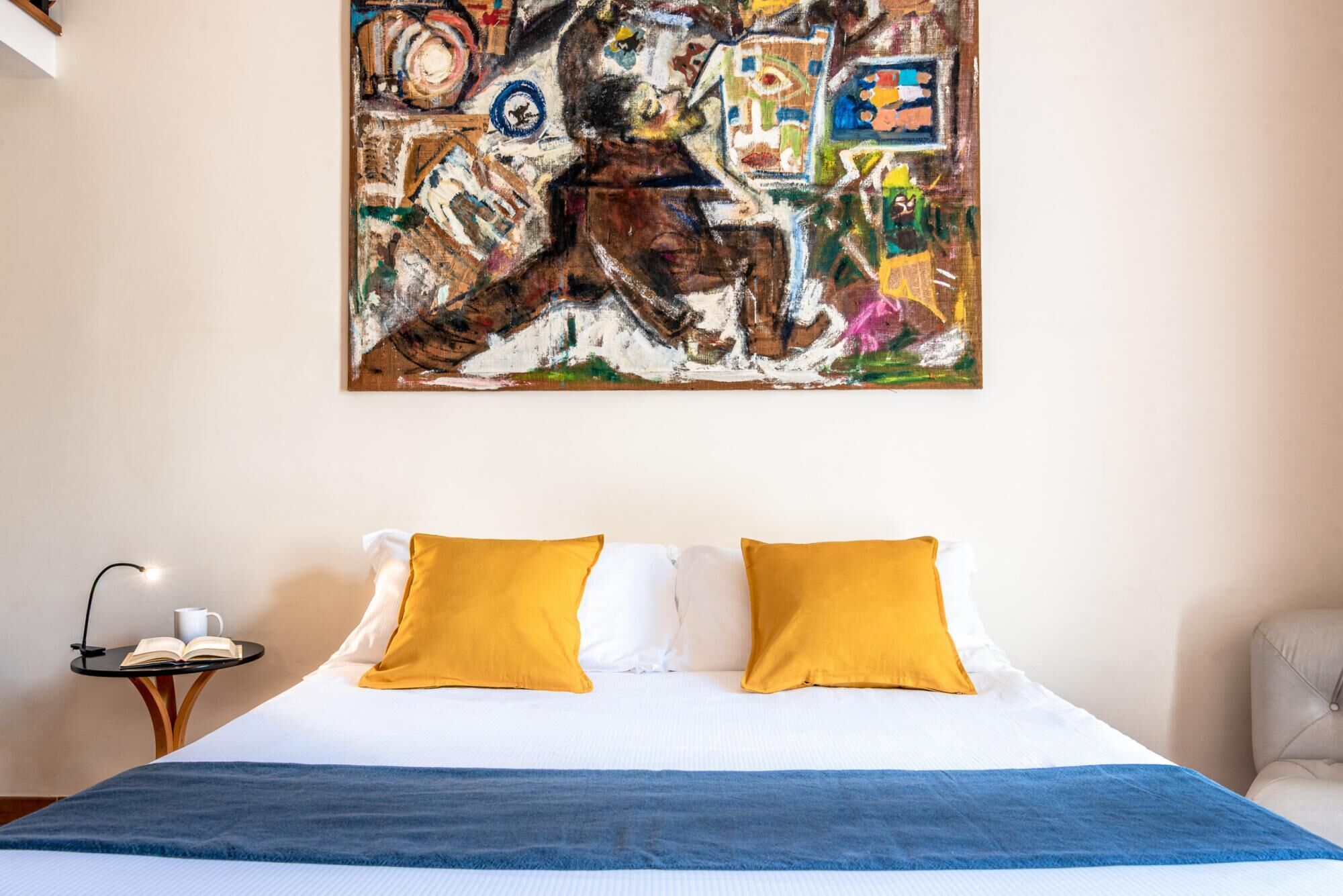 Ein Hotelzimmer mit großem abstrakten Gemälde an der Wand, einem Bett mit weiß-gelben Kissen und einer blauen Decke.