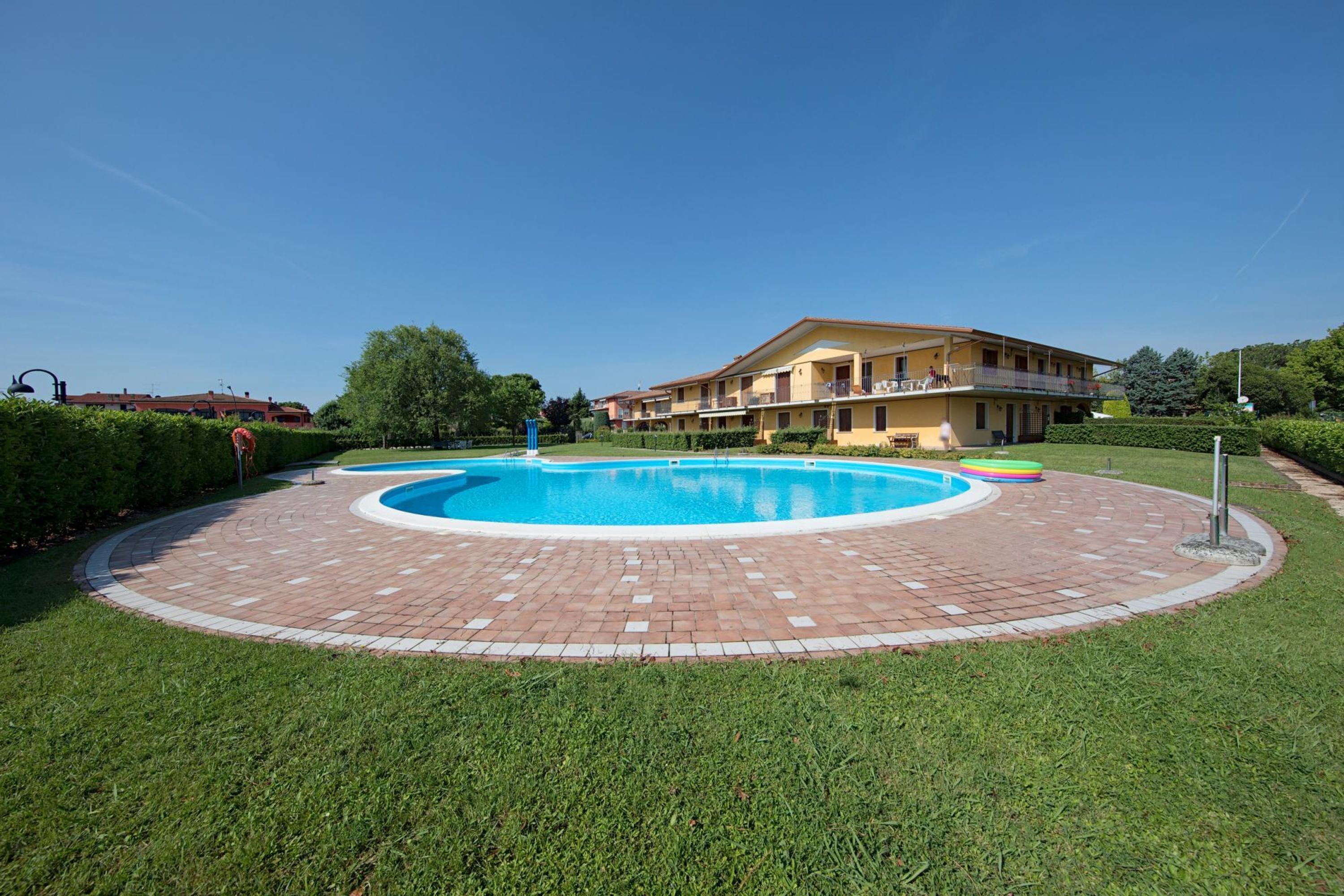 Una piscina circolare con bordo in mattoni, circondata da un prato ben curato, con un edificio con balconi sullo sfondo.