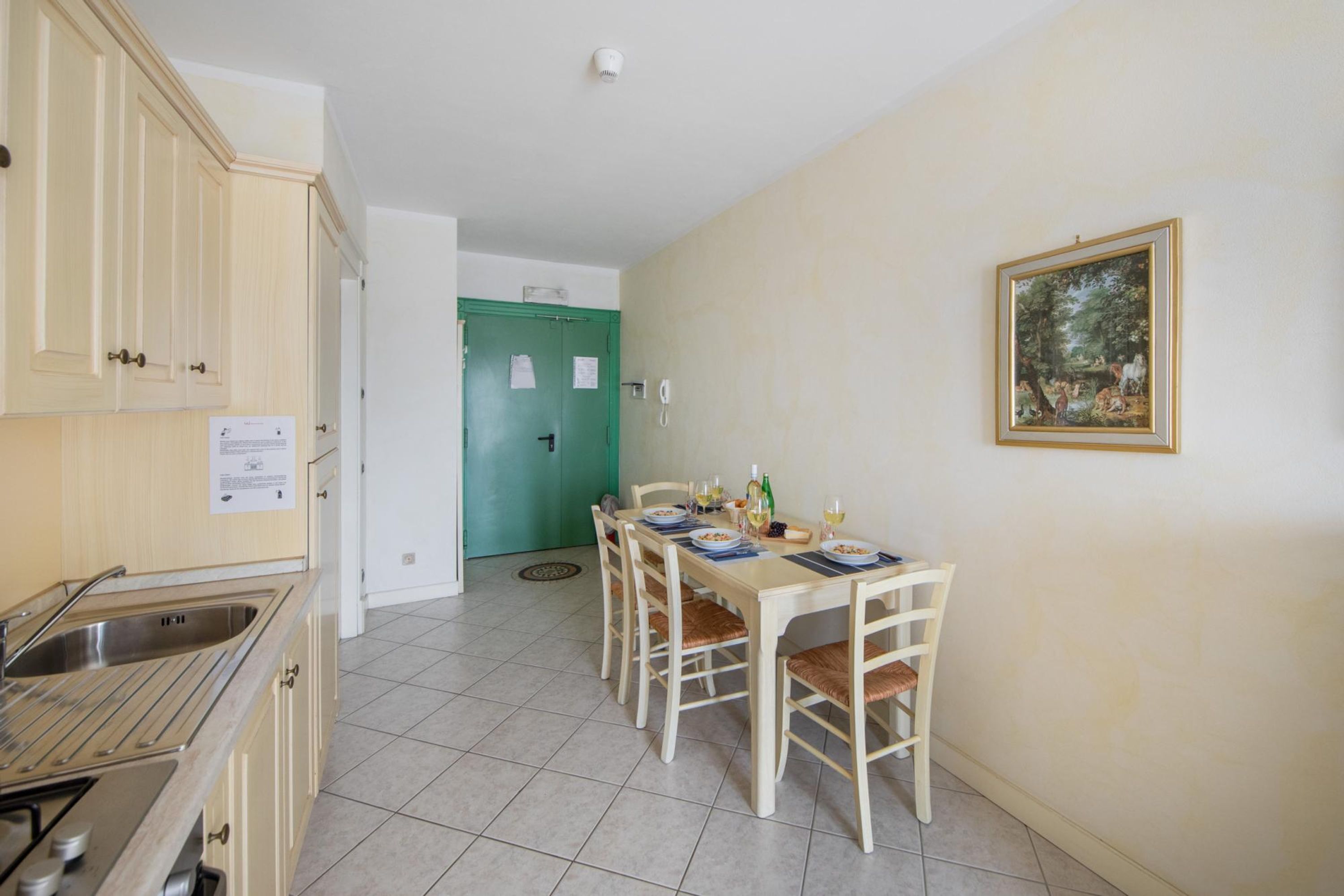 A kitchen with a dining table set for four, a green door, and a framed painting on the wall.