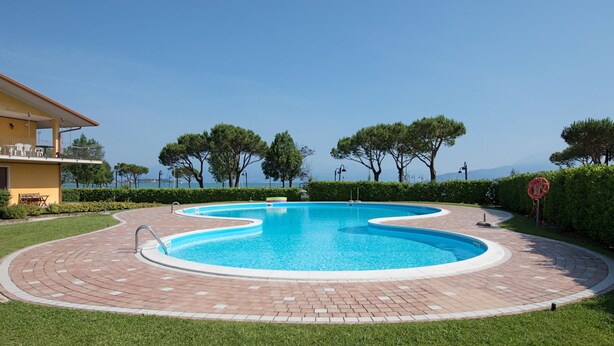 Una piscina circolare con un vialetto in mattoni intorno, un edificio giallo alla sinistra e alberi sullo sfondo.