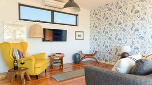A living room with a yellow armchair, a grey sofa, a wooden coffee table, a small side table, a vase, and a television mounted on the wall. - Daydream Cottage (Daylesford)