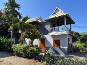 A two-story building with a balcony, surrounded by greenery and palm trees.