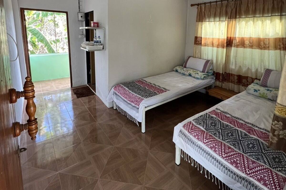 A room with two beds, a ceiling fan, a window with curtains, and a door with a wooden handle.
