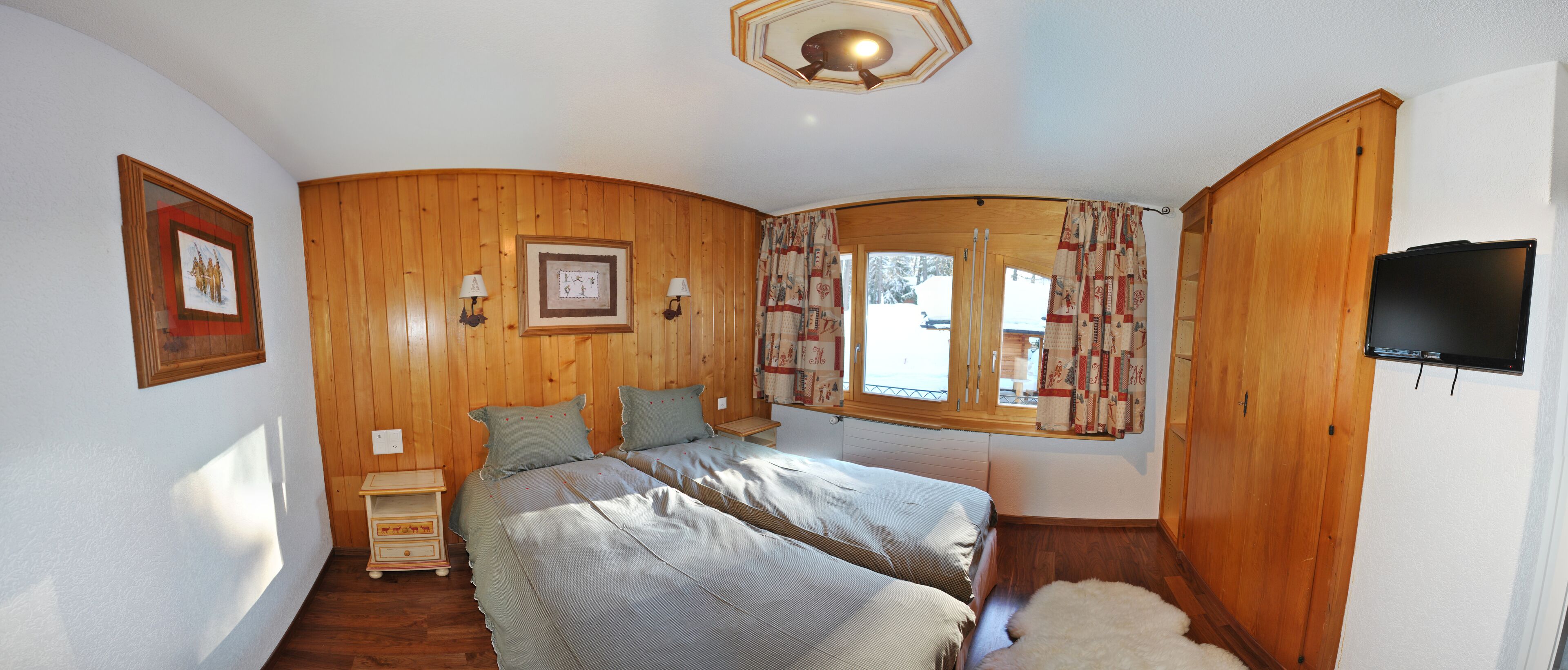 Een slaapkamer met twee bedden, houten wanden, een raam met gordijnen, een nachtkastje en een aan de muur bevestigde televisie.