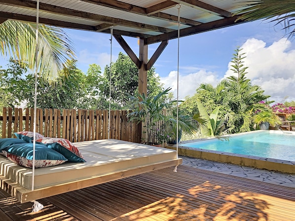 Un hamac avec des coussins, une terrasse en bois, une piscine et une végétation luxuriante.