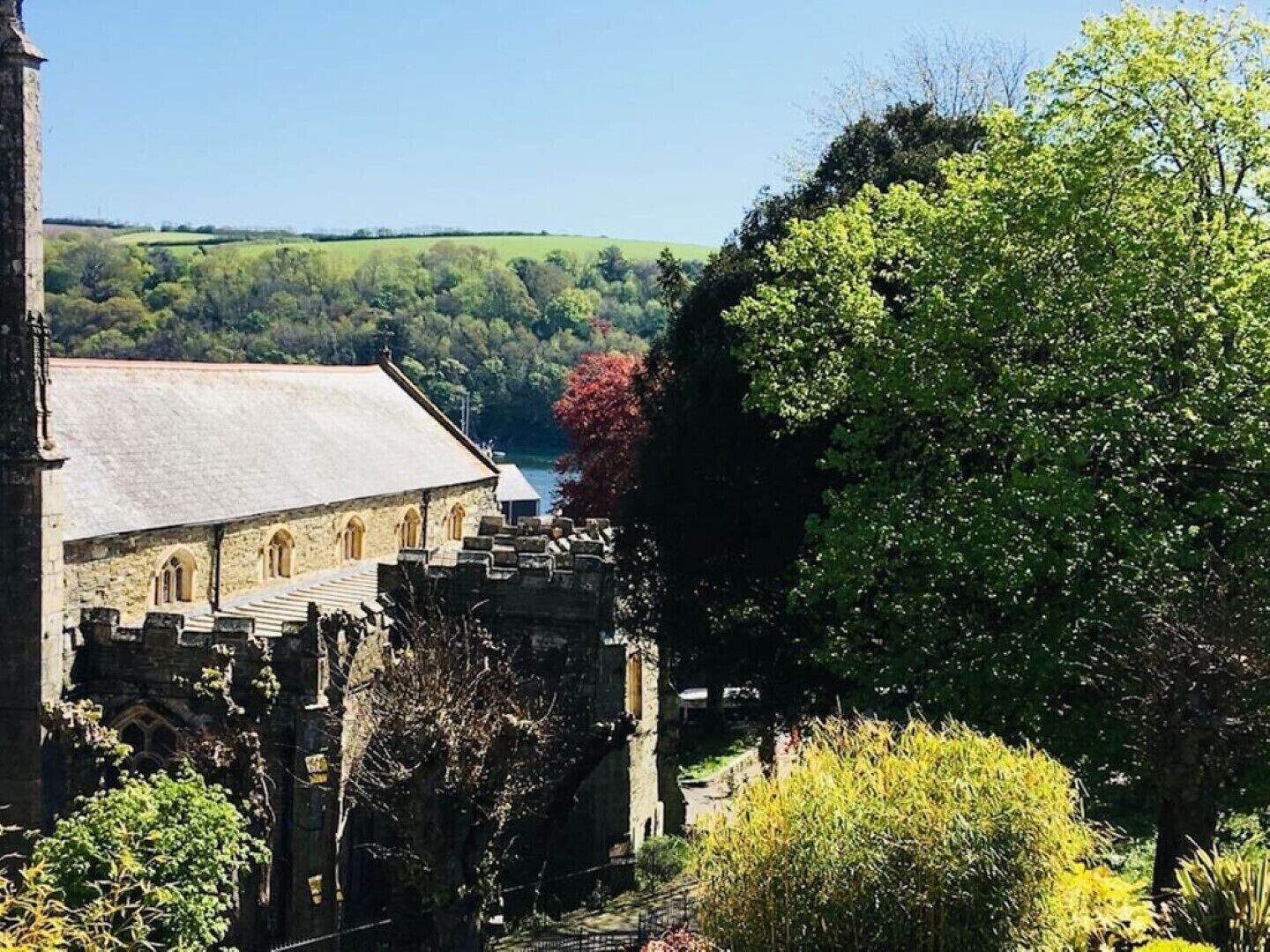 A historic stone building with a large arched entrance and a steeply pitched roof, surrounded by lush greenery and a clear blue sky.