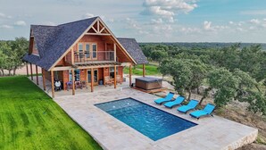 A two-story log cabin with a balcony, a hot tub, and a swimming pool with lounge chairs. - Exquisite Hill Country Cottage-Giraffe and wildlife experiences (Lometa)