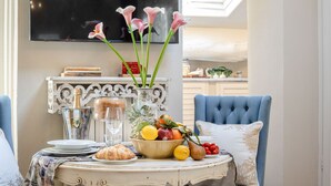 Una mesa de comedor con un frutero, una botella y un croissant. Se ven sillas y un televisor al fondo.