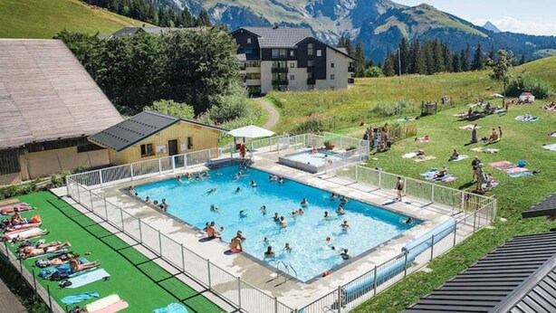 A swimming pool surrounded by a fenced area with people sunbathing and swimming. In the background, there is a large building with a sloped roof and a mountainous landscape. - RDC studio on the slopes, mountain views (Manigod)