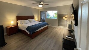 Een slaapkamer met een bed, nachtkastjes, een plafondventilator, een airconditioningapparaat, een televisie en een raam met jaloezieën.
