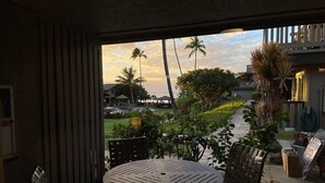 Uitzicht vanuit een raam van een kamer met een ronde tafel en stoelen, een balkon met een leuning, en een aangelegde tuin met palmboompjes en een gebouw op de achtergrond.