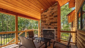 A wooden deck with a stone fireplace, outdoor seating, and a view of the forest. - Spacious Mountain Lodge w/ Hot Tub & Fire Pit (McHenry)