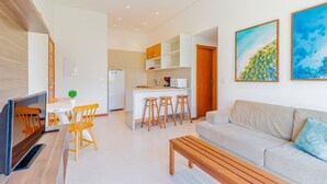 Ein modernes Wohnzimmer mit einer Couch, einem Holztisch und einem Fernseher. Es gibt eine Küchenzeile mit Kühlschrank, Mikrowelle und Barhocker.