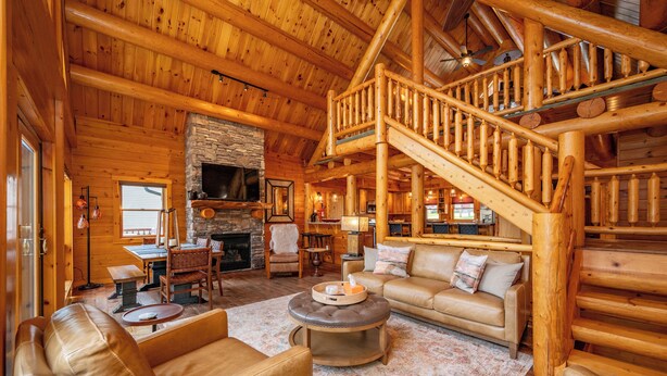 A log cabin interior with a stone fireplace, wooden staircase, and leather furniture. - Stylish Lakefront Escape w/ Hot Tub, Bar & Dock (Oakland)