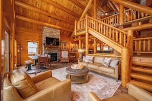 A log cabin interior with a stone fireplace, wooden staircase, and leather furniture.