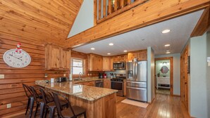A spacious kitchen with wooden cabinetry, a granite island with bar stools, stainless steel appliances, and a clock on the wall. - Woodsy Retreat Near Wisp w/ Golf & Hot Tub (McHenry)