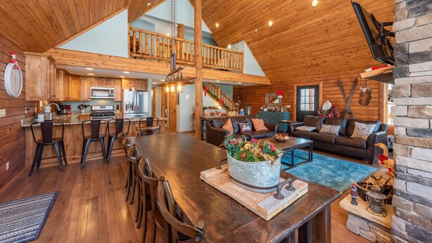 A spacious loft-style living area with a high ceiling, wooden beams, a dining table with chairs, a sofa, and a kitchen in the background. - Woodsy Retreat Near Wisp w/ Golf & Hot Tub (McHenry)