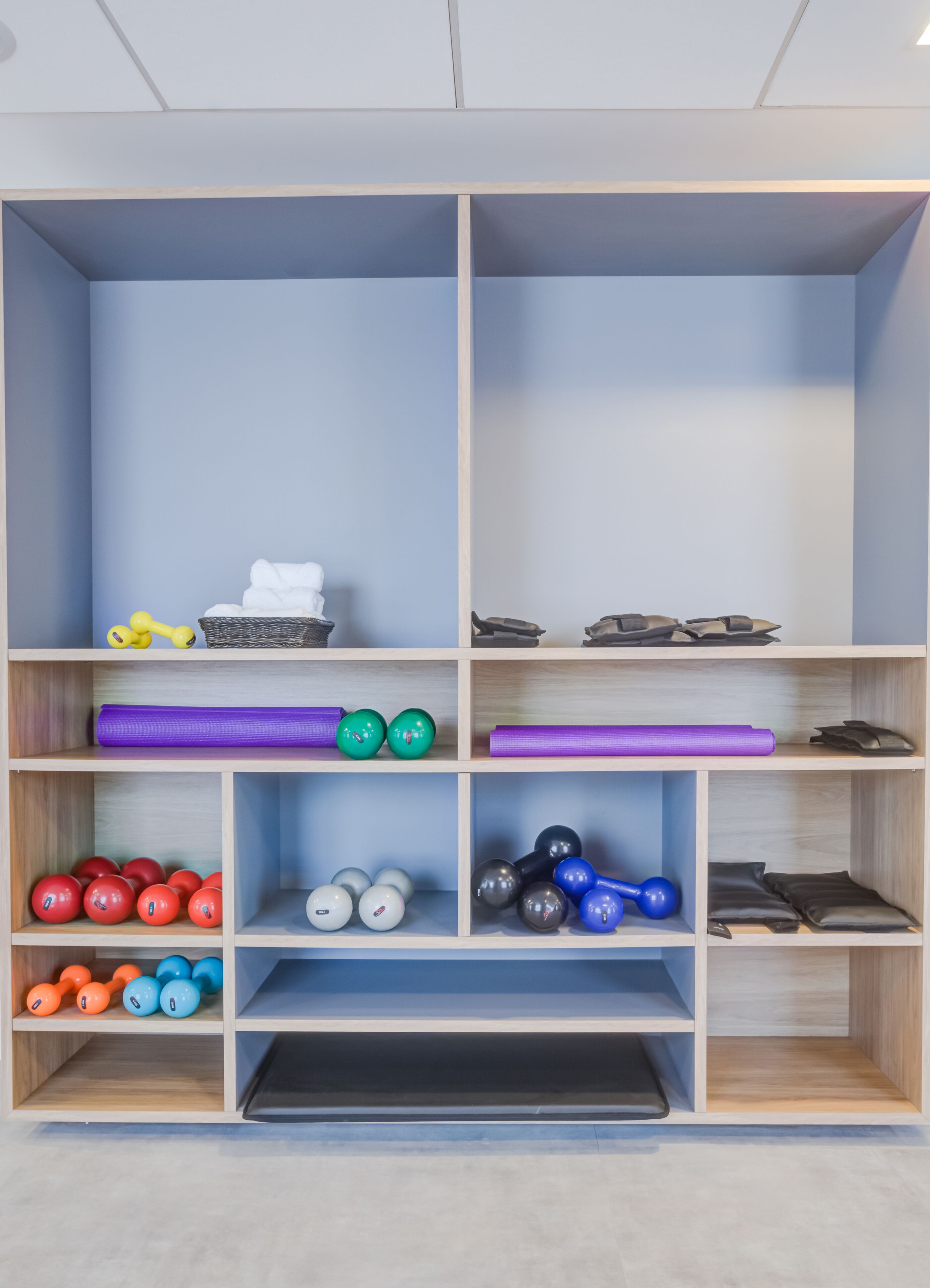 A storage unit with multiple shelves, some containing exercise equipment like dumbbells and yoga mats, and others holding items like towels and a basket.
