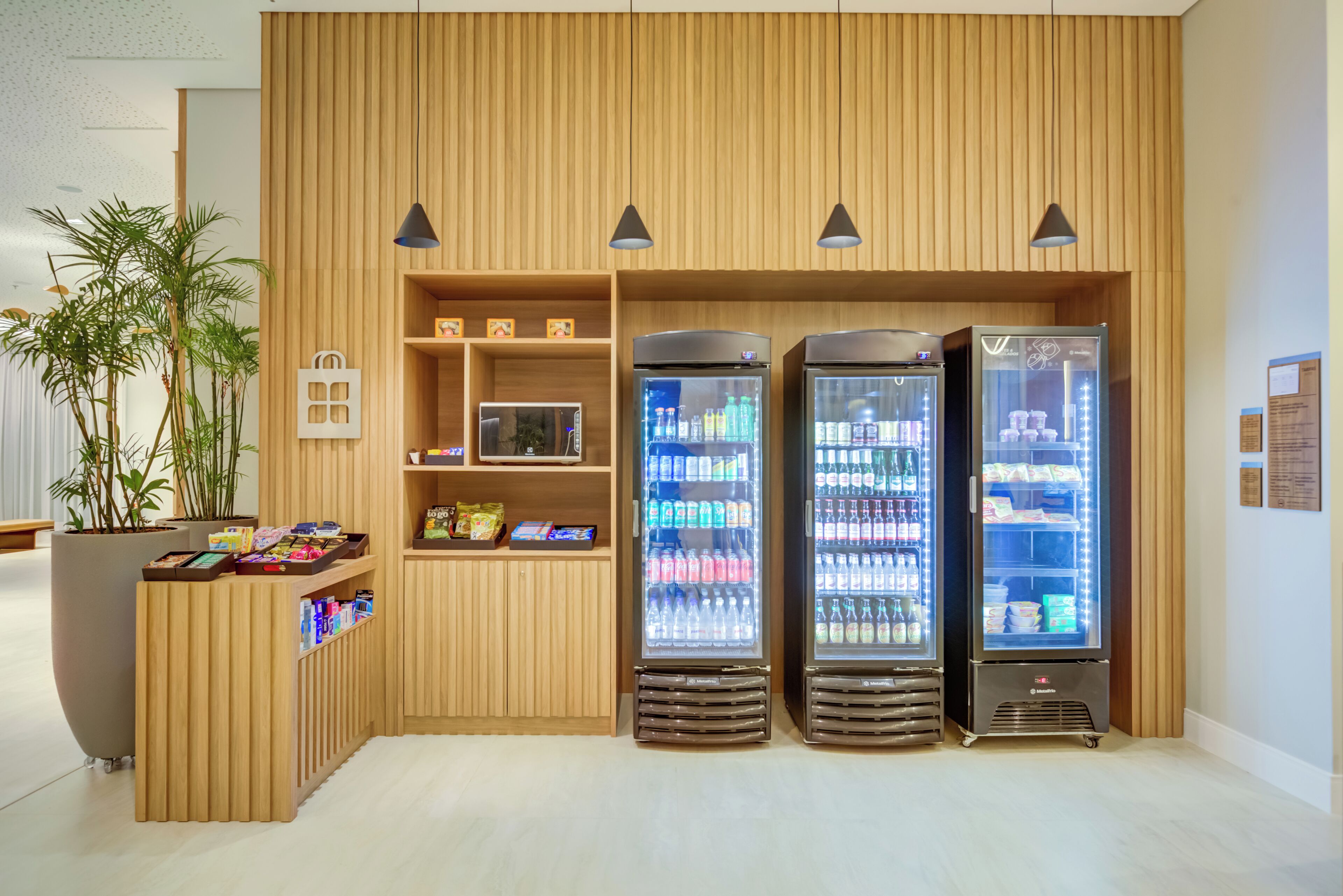 Un quiosco moderno con tres dispensadores refrigerados para bebidas, una planta en maceta y un mostrador de madera con estantes.